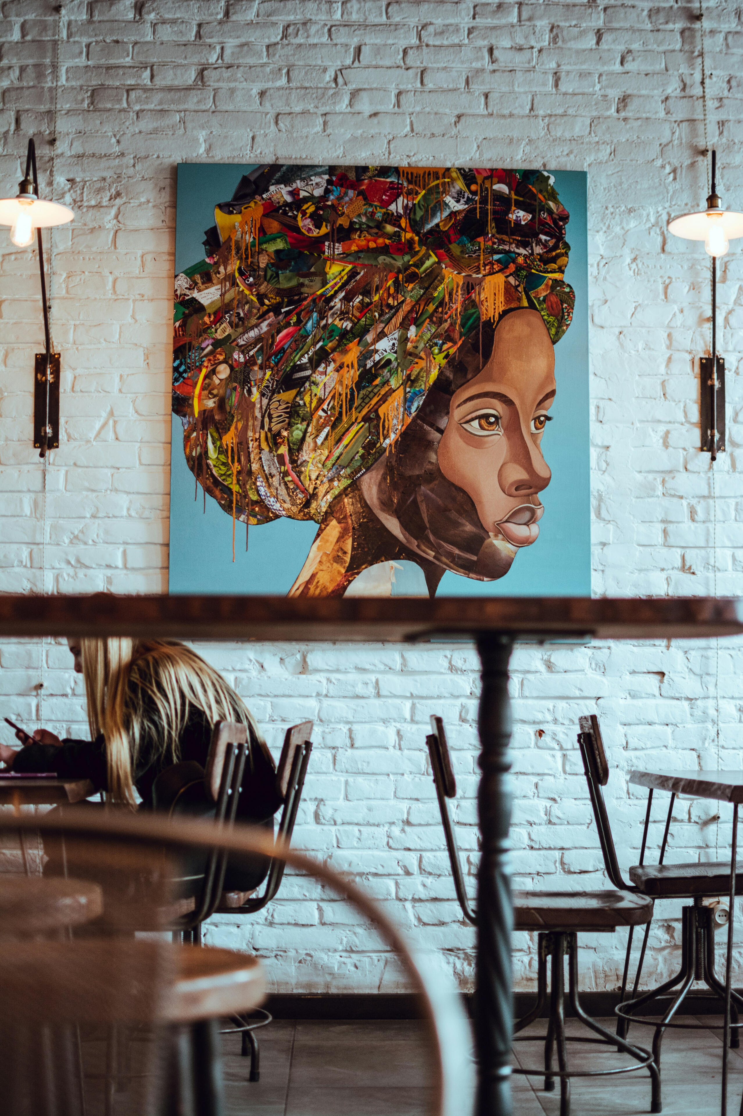 A multi-colored portrait of a woman hanging on a white brick wall in a cafe. There is a woman sitting at a table in front of the painting.